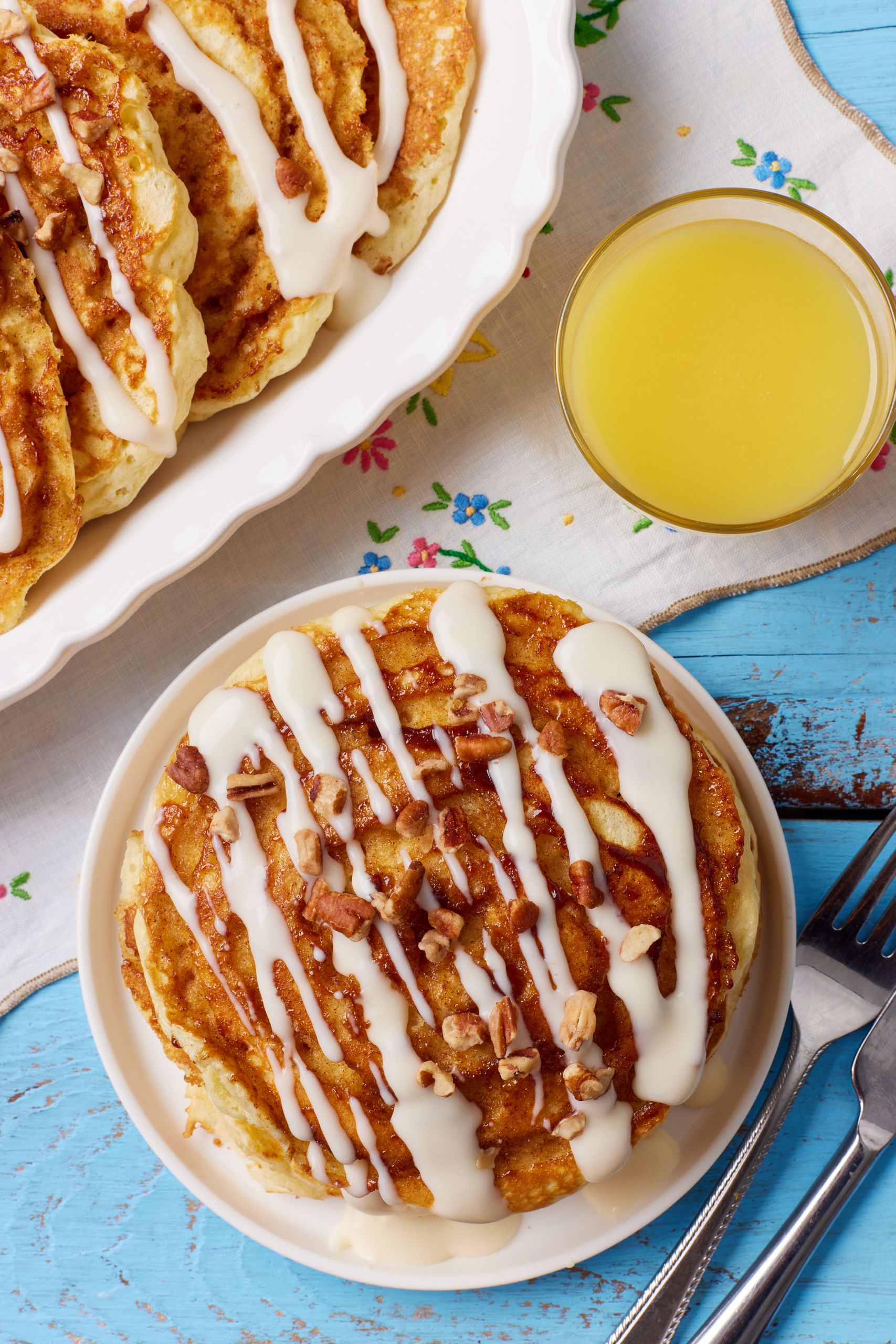 Cinnamon Roll Pancakes11 A top-down view of golden fluffy cinnamon bun pancakes served with nuts and cream cheese glaze, with orange juice on the side.