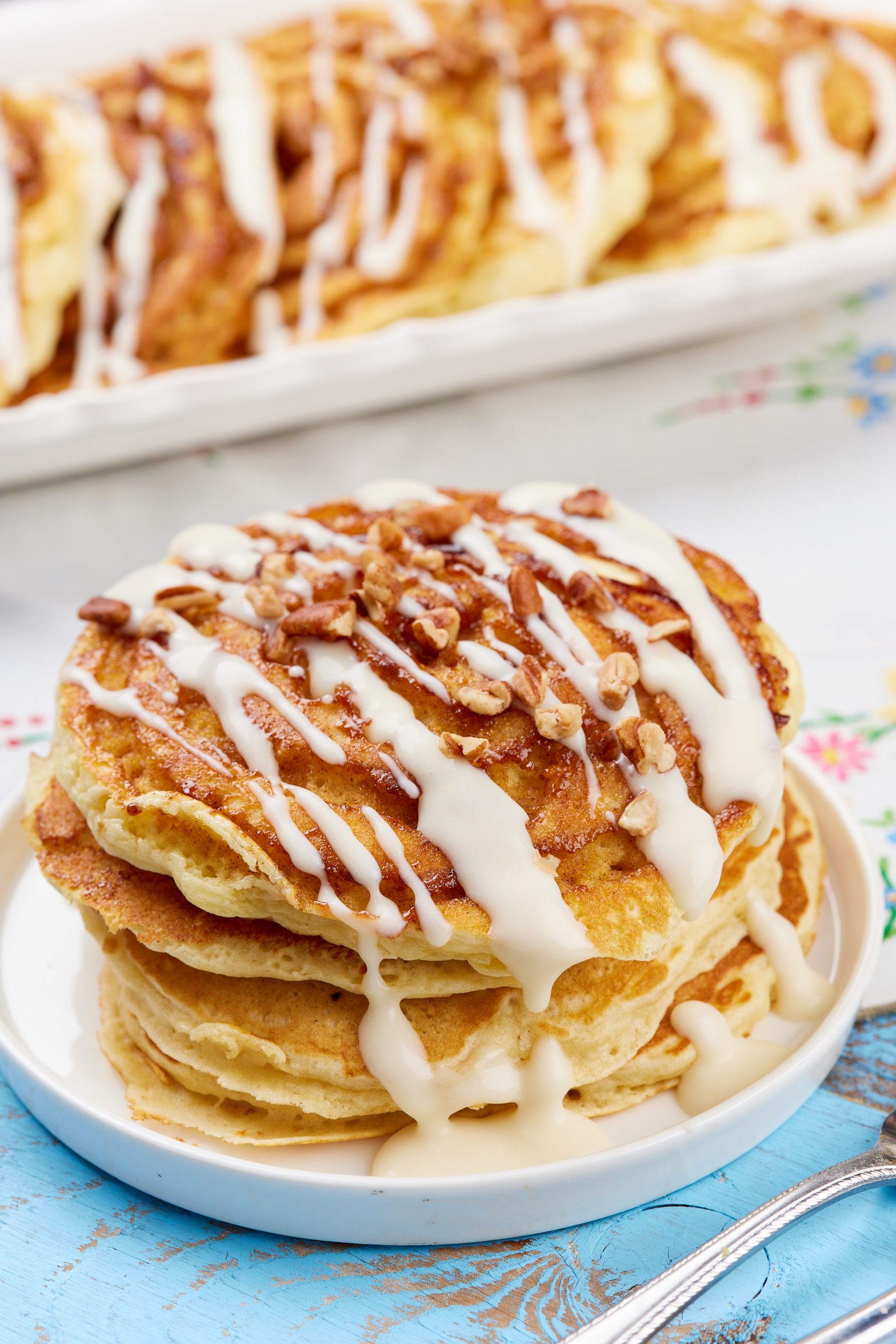 Cinnamon Roll Pancakes12 A stack of golden fluffy Cinnamon Roll Pancakes topped with toasted pecans and drizzled with cream cheese glaze.