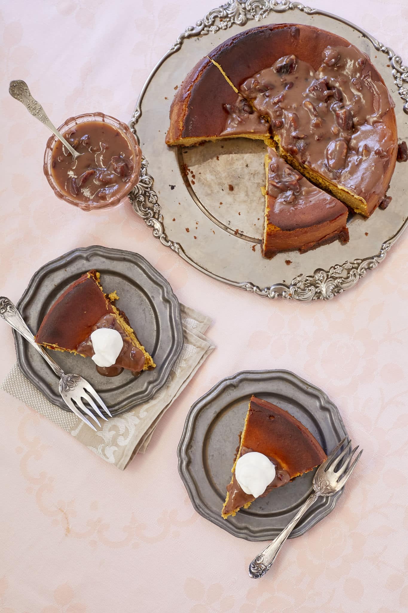 Homemade pumpkin cheesecake with pecan praline sauce is served on a platter. Two slices are dolloped in homemade whipped cream.