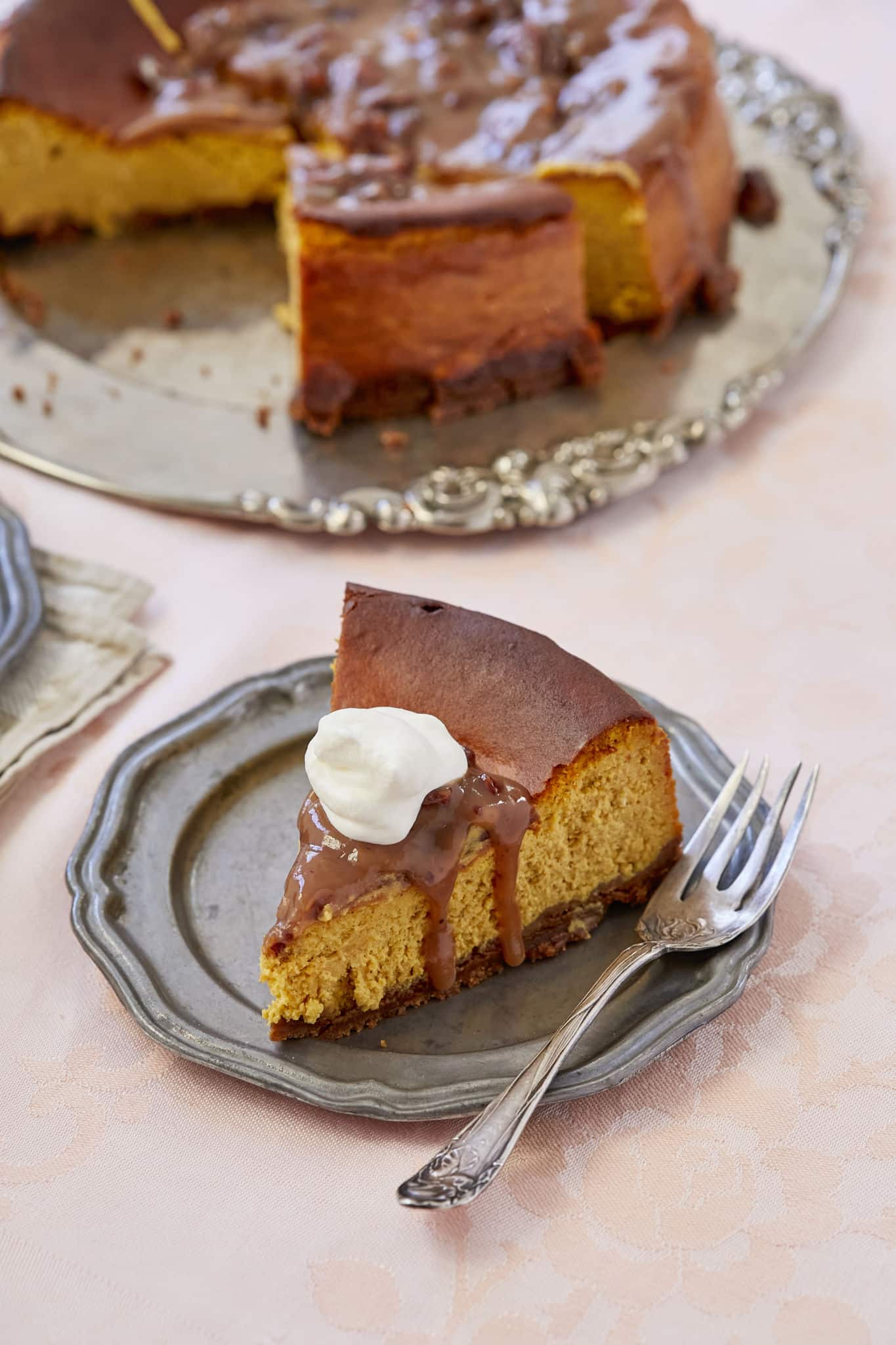 Pumpkin cheesecake with pecan praline sauce is served on a silver plater with a dollop of whipped cream.