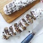 One chocolate salami is on the chopping board and the others sliced not is on the counter. The charming marbled interior of this chocolate salami surprises you at first sight when sliced.  This mixture is shaped into a log, resembling a salami, and is often rolled in powdered sugar or cocoa powder to simulate the look of cured meat.