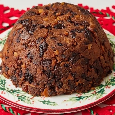 Traditional Christmas pudding packed with dried fruit and candied peel, served on a festive Christmas plate with custard and gold spoons.