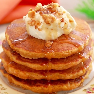 Carrot Cake Pancakes are in vibrant orange color, and stacked together. Topped with cream cheese frosting and toasted walnuts.