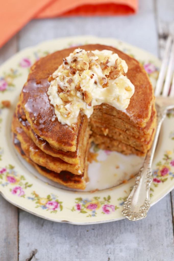 Carrot Cake Pancakes are in vibrant orange color, and stacked together. Topped with cream cheese frosting and toasted walnuts.