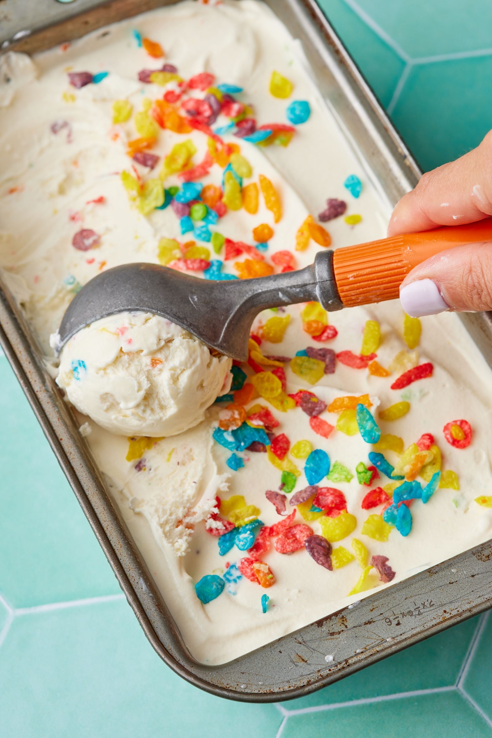 A top-down view of cereal milk ice cream in a loaf tin, sprinkled with Fruity pebble. One scoop is being served. 