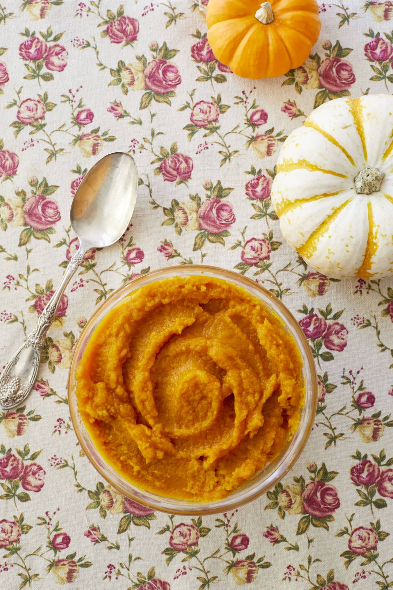 Homemade Pumpkin puree is in a glass bowl in a vibrant orange color and with a smooth moist consistency. A silver spoon is on the left and an orange sugar pumpkin and a white pumpkin are on the right side.