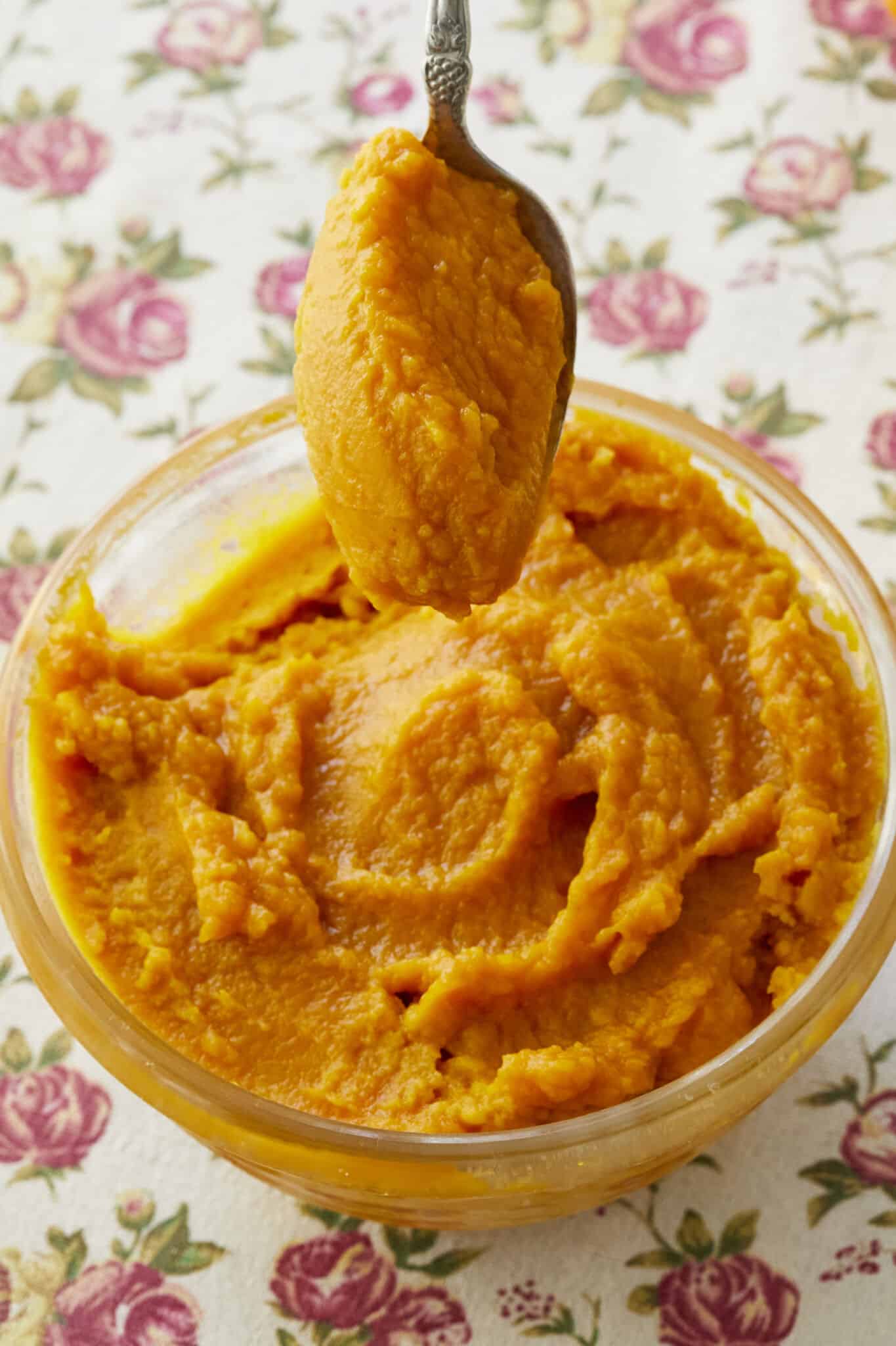 A spoonful of Homemade Pumpkin puree is being lifted from the glass bowl. It's in a vibrant orange color and has a smooth moist consistency. 