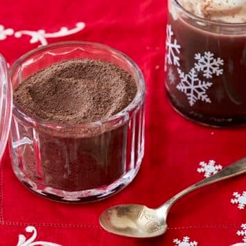 Homemade Hot Chocolate Mix in a glass jar. A mug of hot chocolate made from this is next to it with whipped cream on top.