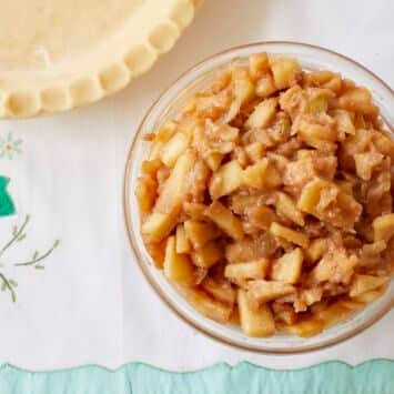 Crock Pot Apple Pie Filling is in a glass bowl, ready for the assembled pie crust next to it. The filling has tender slow cooker apples  in chunks covered with cinnamon and sticky gooey sauce.