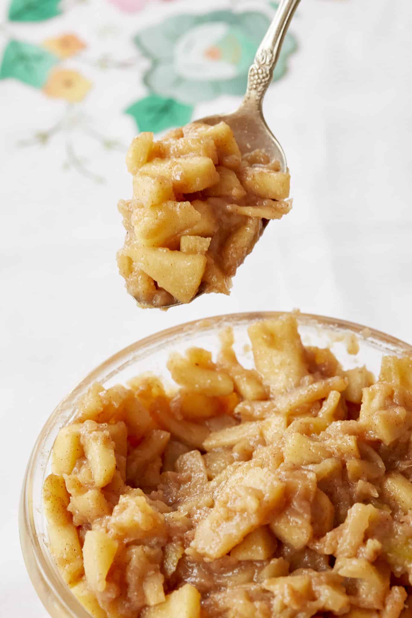 A spoon of apple pie filling is scooped from the glass bowl. It has chunks of soft apples covered with cinnamon and sticky sweet sauce. 
