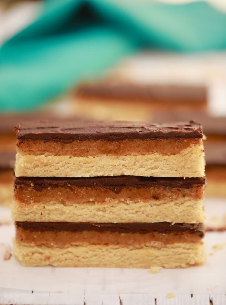 A homemade Twix recipe that uses healthier ingredients is photographed. Three homemade Twix bars are stacked on each other with a layer of cookie, caramel, and chocolate.