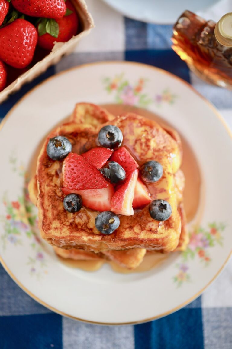 Homemade French Toast is stacked on a dish, topped with fresh strawberries and blueberries, and covered in maple syrup.