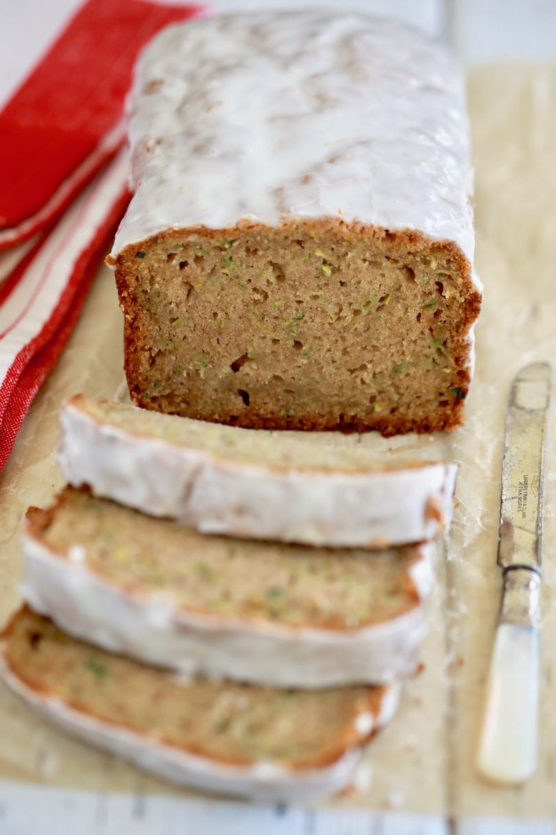 A loaf of glazed zucchini quick bread is sliced in the middle, displaying a tight crumb and bits of the zucchini throughout.