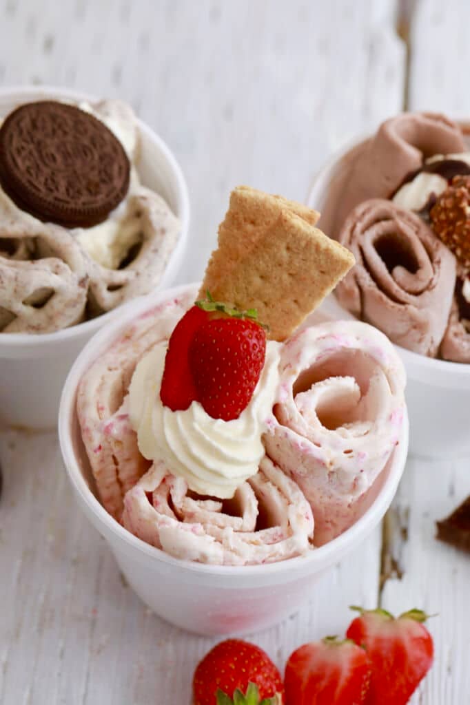3 flavor combinations of Rolled Ice Cream. The Strawberry flavor is in the front with whipped cream, fresh strawberry and graham crackers