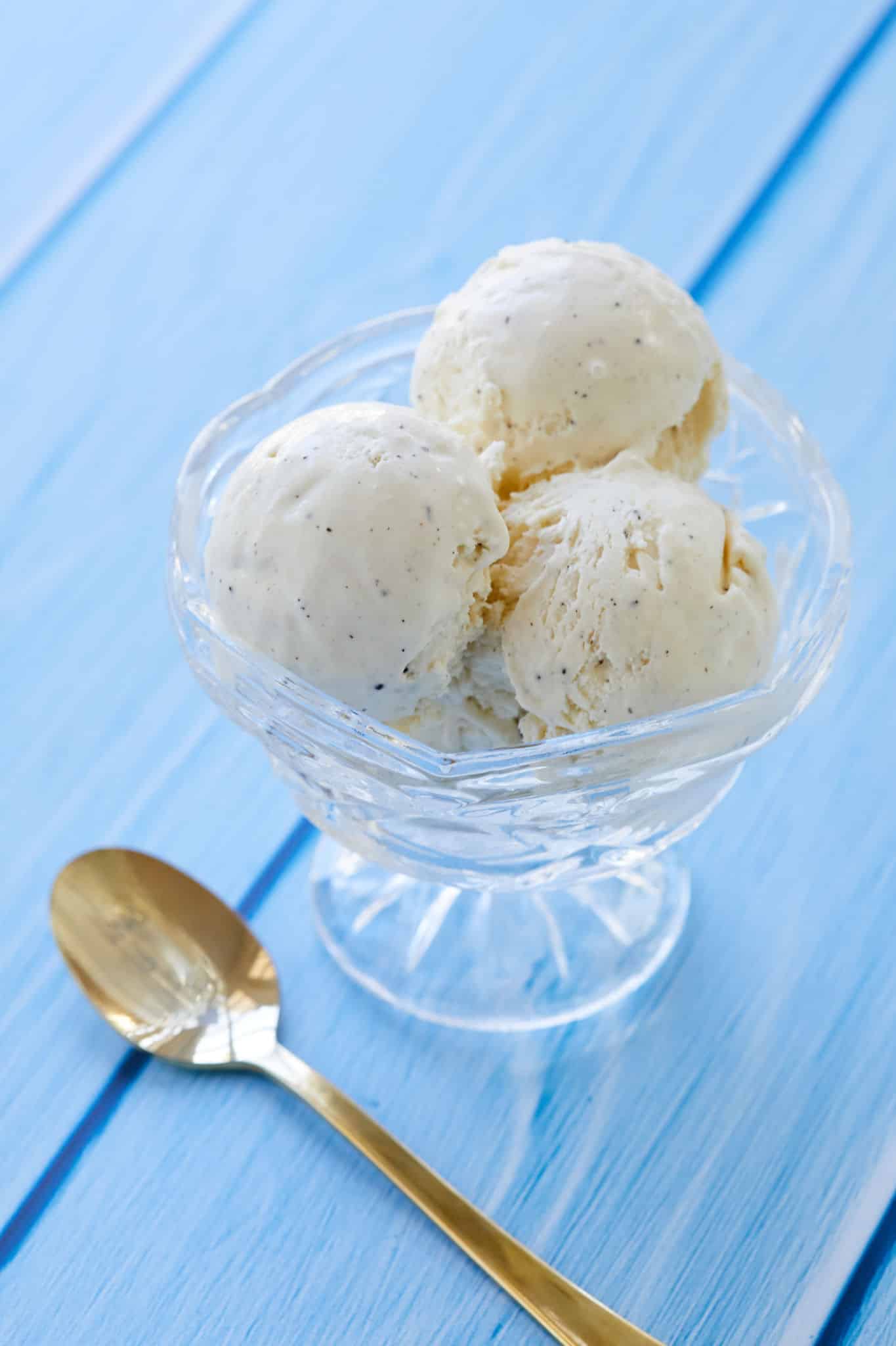 Creamy homemade vanilla ice cream with vanilla seeds dispersed throughout, served in a glass bowl with a golden spoon on the left. 