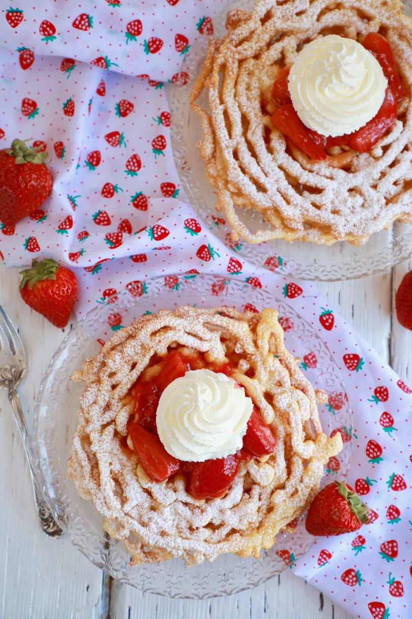 Top-down view of two funnel cakes topped with strawberries and whipped cream.