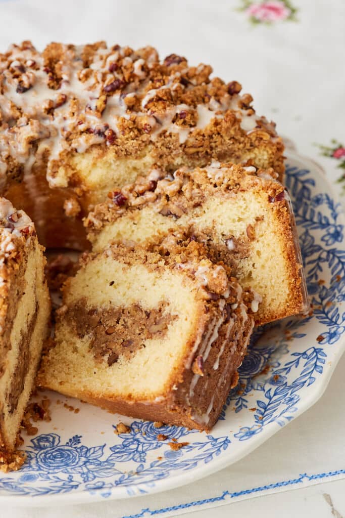 A close-up of a slice of coffee cake with a moist, tender crumb, a cinnamon-nut filling, and a generous crumb topping drizzled with glaze, served on a blue-and-white floral plate.