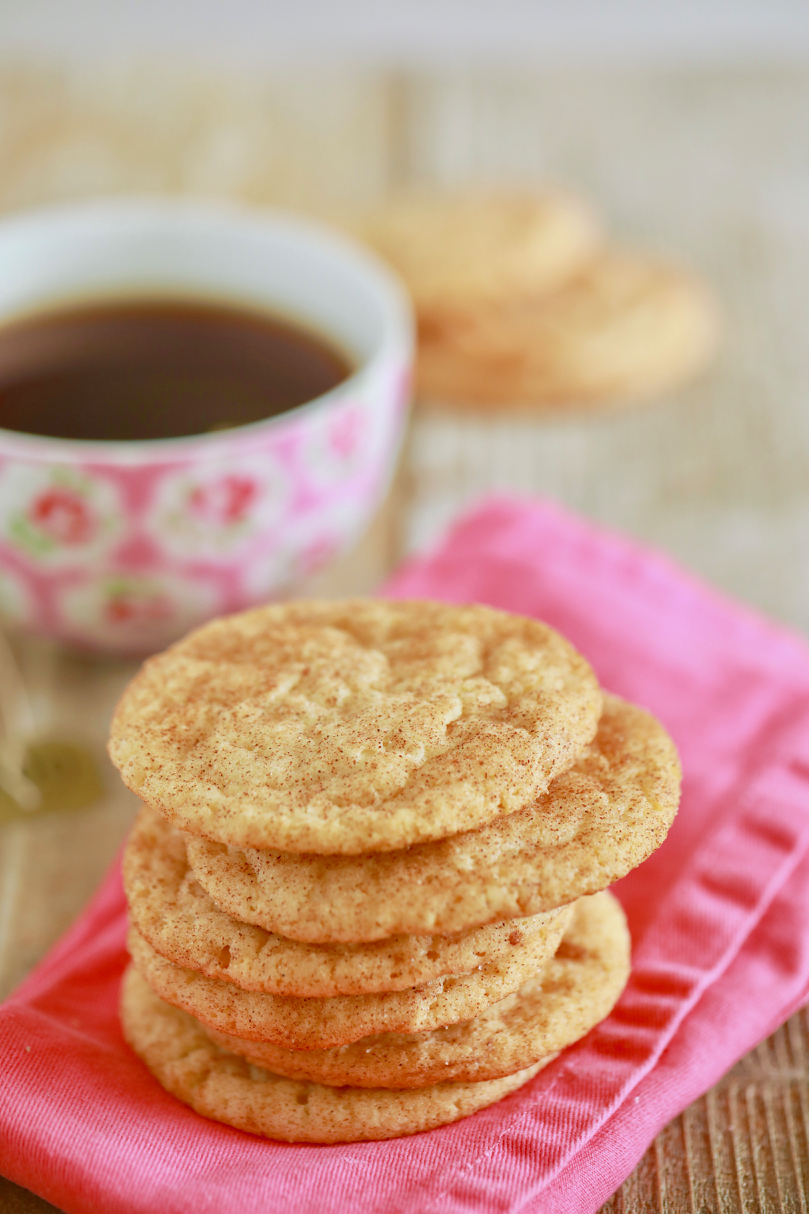 A stack of snickerdoodle cookies rest on a pink napkin in front of a cup of tea in a floral teacup. 