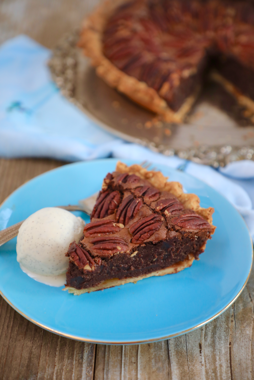 A slice of pecan fudge pie is served on a blue plate, the filling is a deep chocolate brown with pecans on top. It is served with a side of vanilla ice cream.