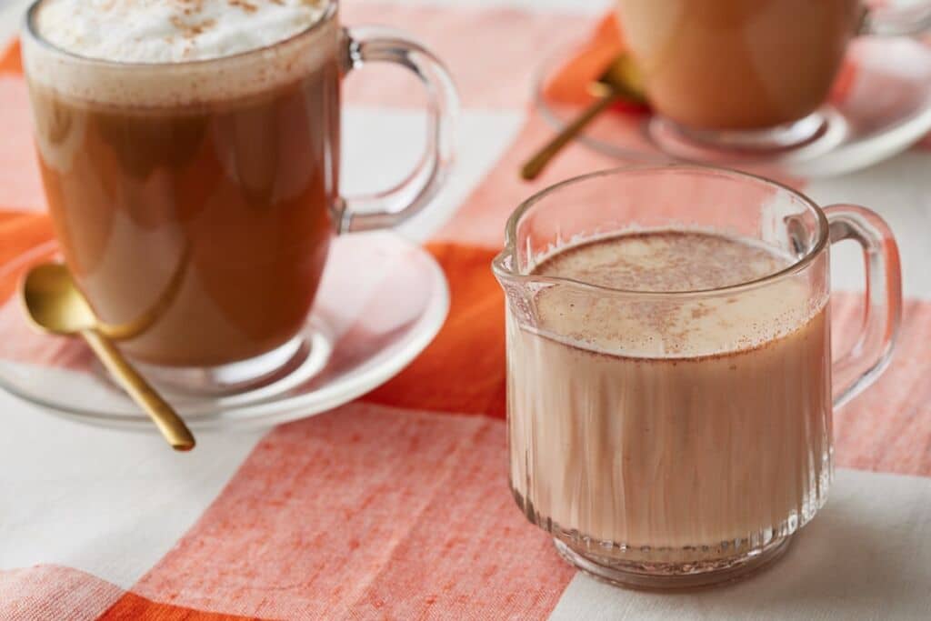 Pumpkin spice coffee creamer in a glass mug, with whipped cream and cinnamon-topped coffee in the background.