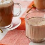 Pumpkin spice coffee creamer in a glass mug, with whipped cream and cinnamon-topped coffee in the background.