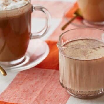 Pumpkin spice coffee creamer in a glass mug, with whipped cream and cinnamon-topped coffee in the background.