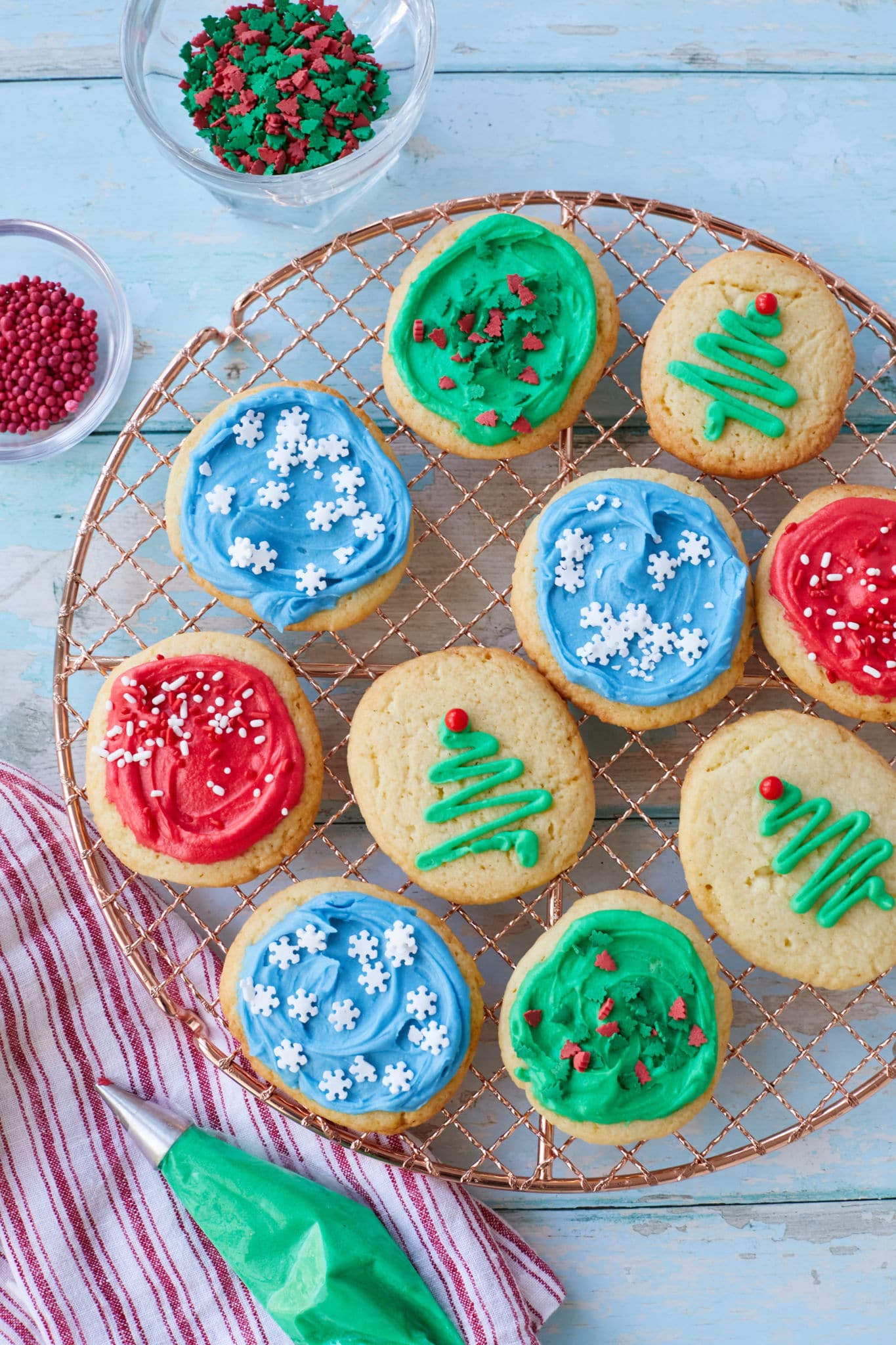 Homemade soft sugar cookies are presented on a golden wire rack. The sugar cookies are decorated in different colored icing and toppings, including red, blue, and green icing, snowflake sprinkles, and Christmas tree decoration.