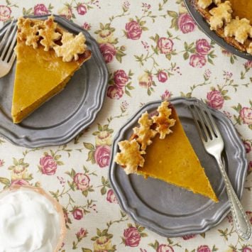 Two slices of homemade pumpkin pie are served on two silver dishes. To the side is a cup of homemade whipped cream.