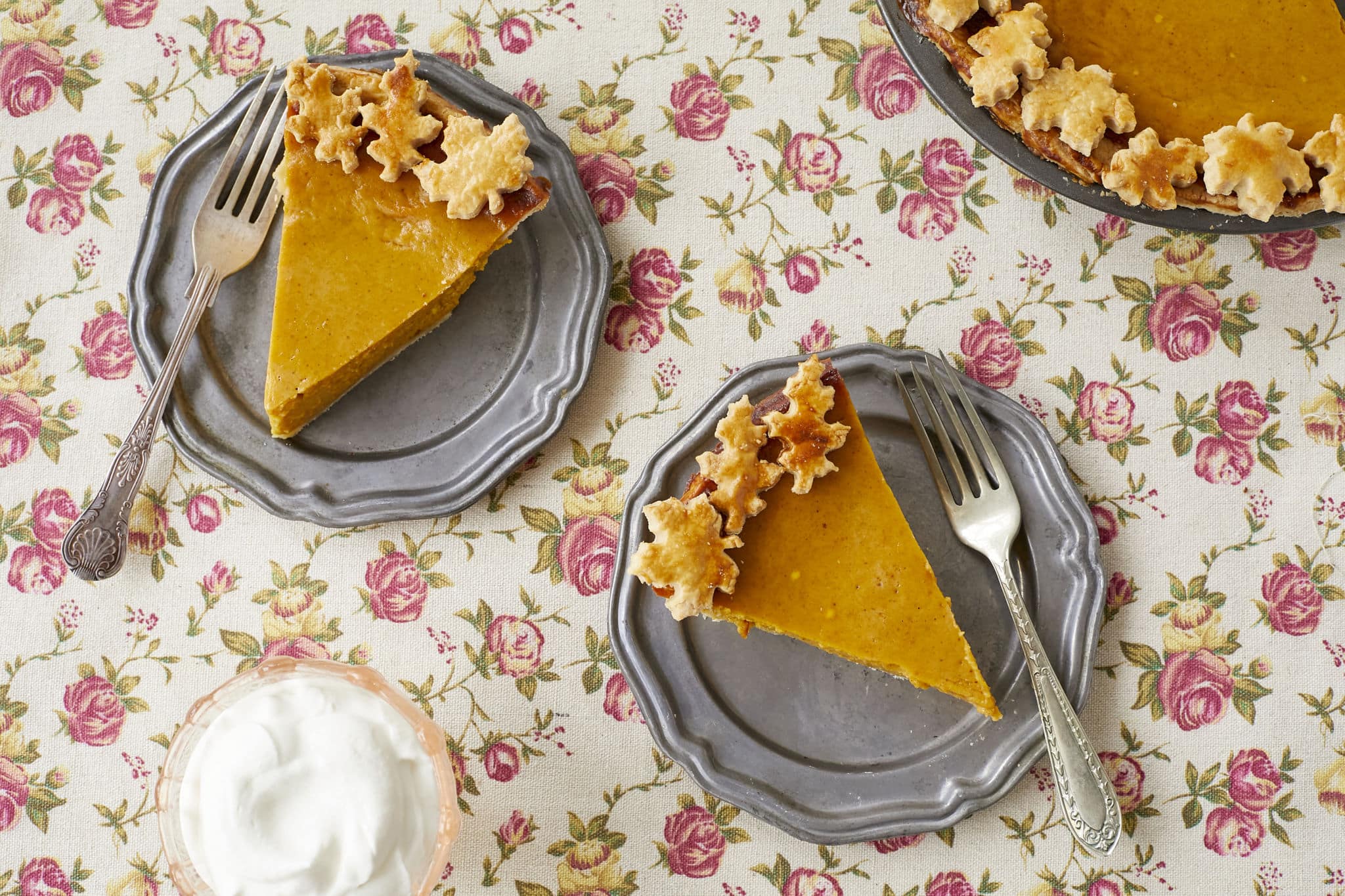 Two slices of homemade pumpkin pie are served on two silver dishes. To the side is a cup of homemade whipped cream.