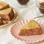 Cranberry Orange Cake is served on a golden-edge platter with two cups of coffee. The slice in front is served on a pink dessert plate, showing the moist interior loaded with cranberries bits and spices. It's covered with glossy lemon glaze which has lemon zest in.