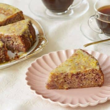 Cranberry Orange Cake is served on a golden-edge platter with two cups of coffee. The slice in front is served on a pink dessert plate, showing the moist interior loaded with cranberries bits and spices. It's covered with glossy lemon glaze which has lemon zest in.
