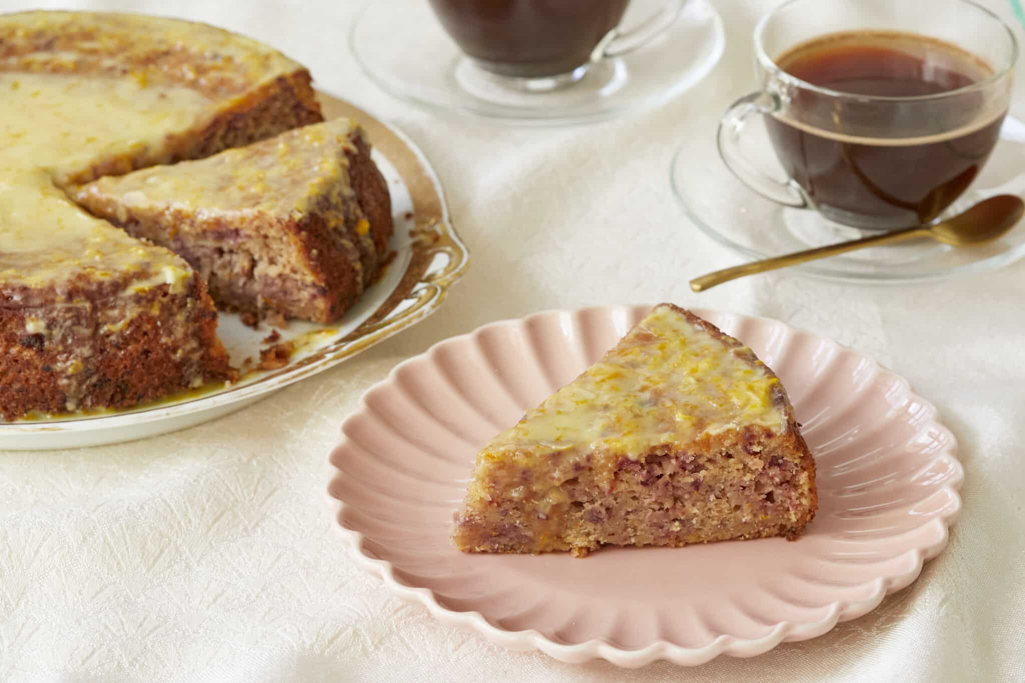Cranberry Orange Cake is served on a golden-edge platter with two cups of coffee. The slice in front is served on a pink dessert plate, showing the moist interior loaded with cranberries bits and spices. It's covered with glossy lemon glaze which has lemon zest in.
