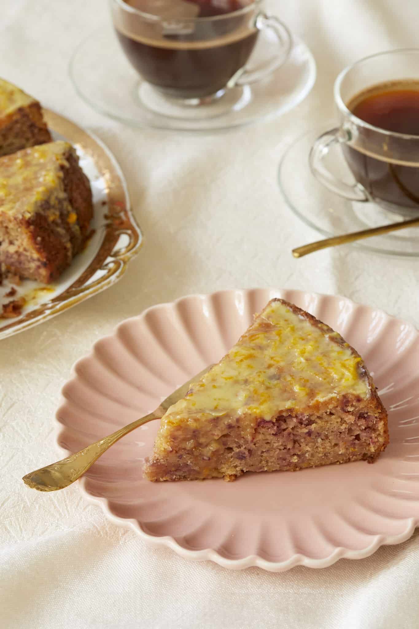 Cranberry Orange Cake is served on a golden-edge platter with two cups of coffee. The slice in front is served on a pink dessert plate, showing the moist interior loaded with cranberries bits and spices. It's covered with glossy lemon glaze which has lemon zest in.