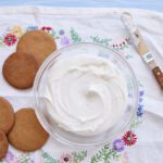 A homemade royal icing is presented in a clear bowl next to five round cookies.