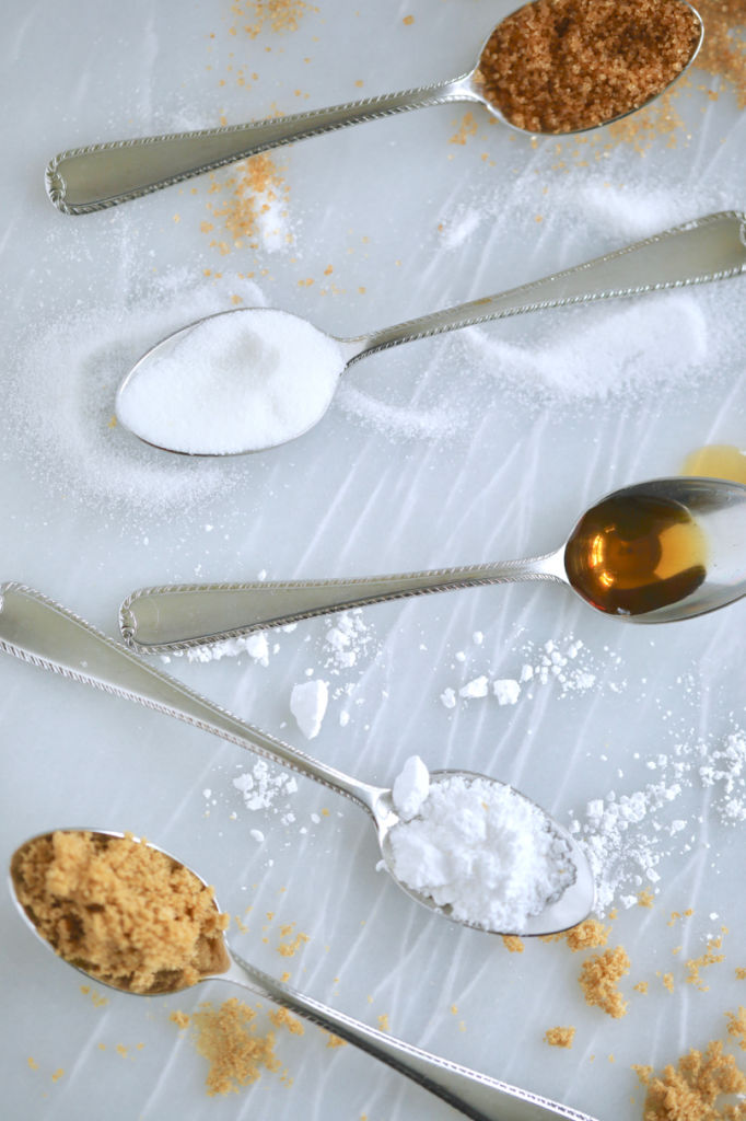 5 Spoons with different sugar substitutes on them, placed haphazardly on a cutting board.