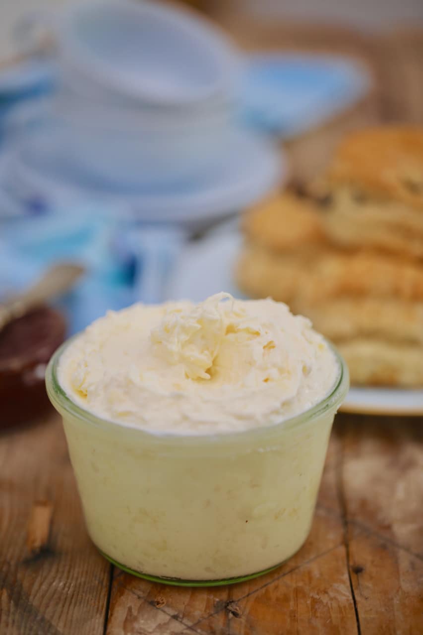 Clotted Cream2 A jar of thick, creamy, yellow Irish Clotted Cream with Irish Scones in the back.