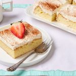 Easy Tres Leches Cake is served with half fresh strawberry and a cup of coffee. The rest of the cake is on the side.