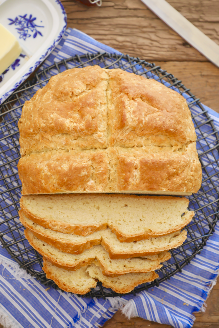 A plump white Irish soda bread sits on a cooling rack over a blue and white napkin. Four slices are cut from the Irish soda bread loaf.