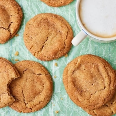 Overhead view of golden-brown snickerdoodle cookies with crackled tops and cinnamon-sugar coating on a mint-green surface, served with a cup of coffee.