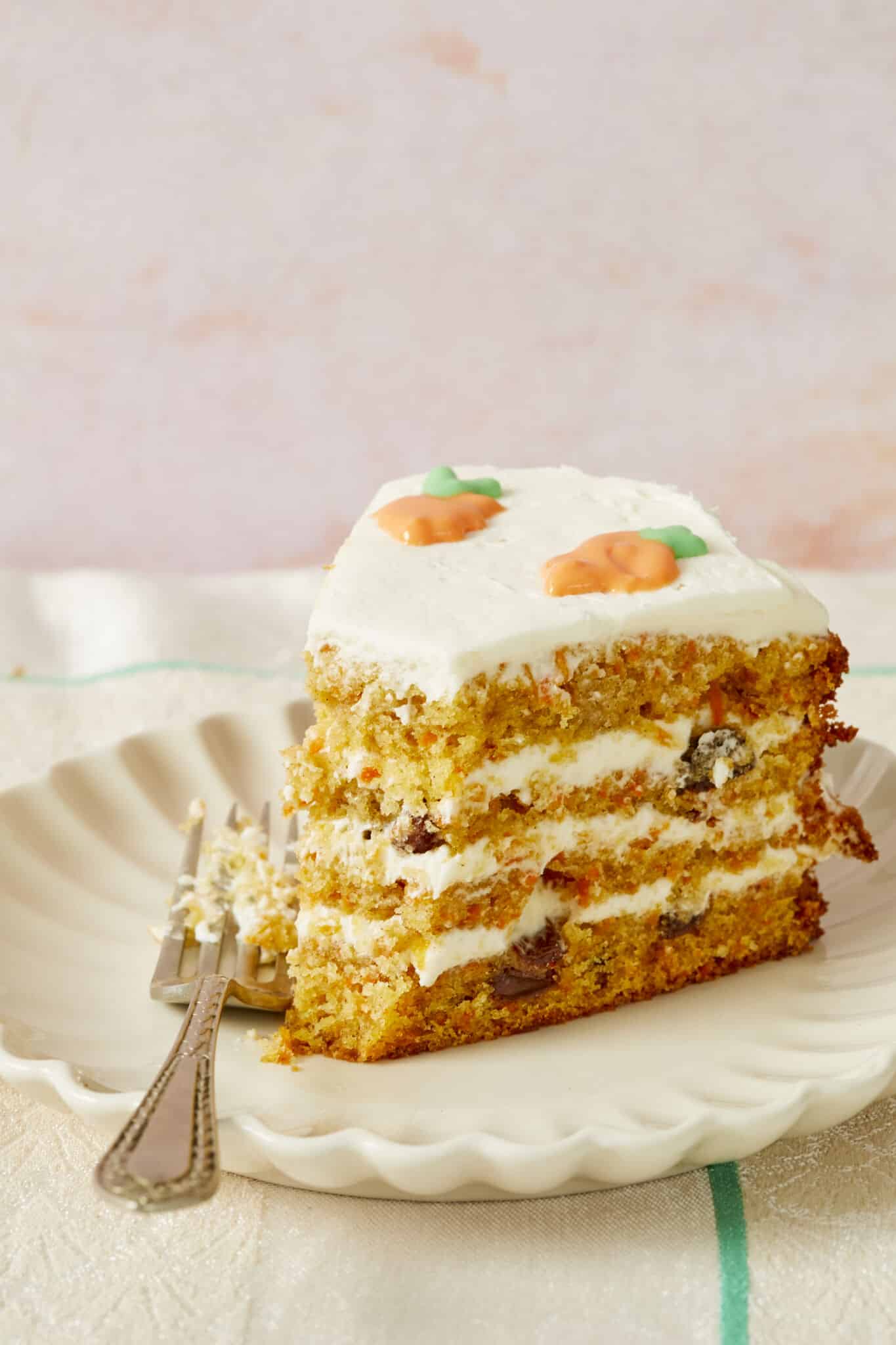 A slice of moist and soft carrot cake full of carrots and raisins, topped with tangy smooth cream cheese frosting, on a plate with one bite taken to enjoy.