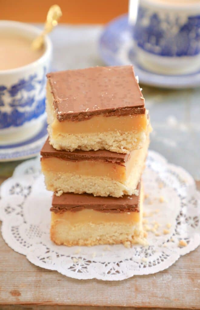 A stack of 3 Millionaire's shortbread squares are severed with tea. They have 3 layers of shortbread, caramel, and chocolate.