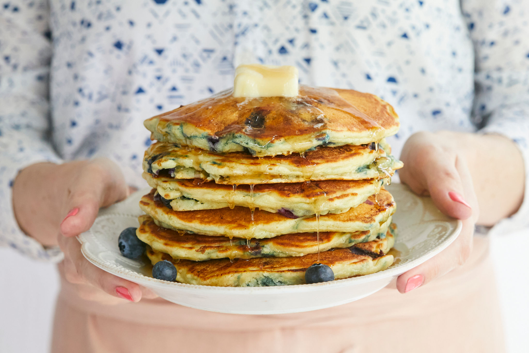 A fresh stack of homemade blueberry pancakes, topped with butter, syrup, and being held on a plate by Gemma Stafford.