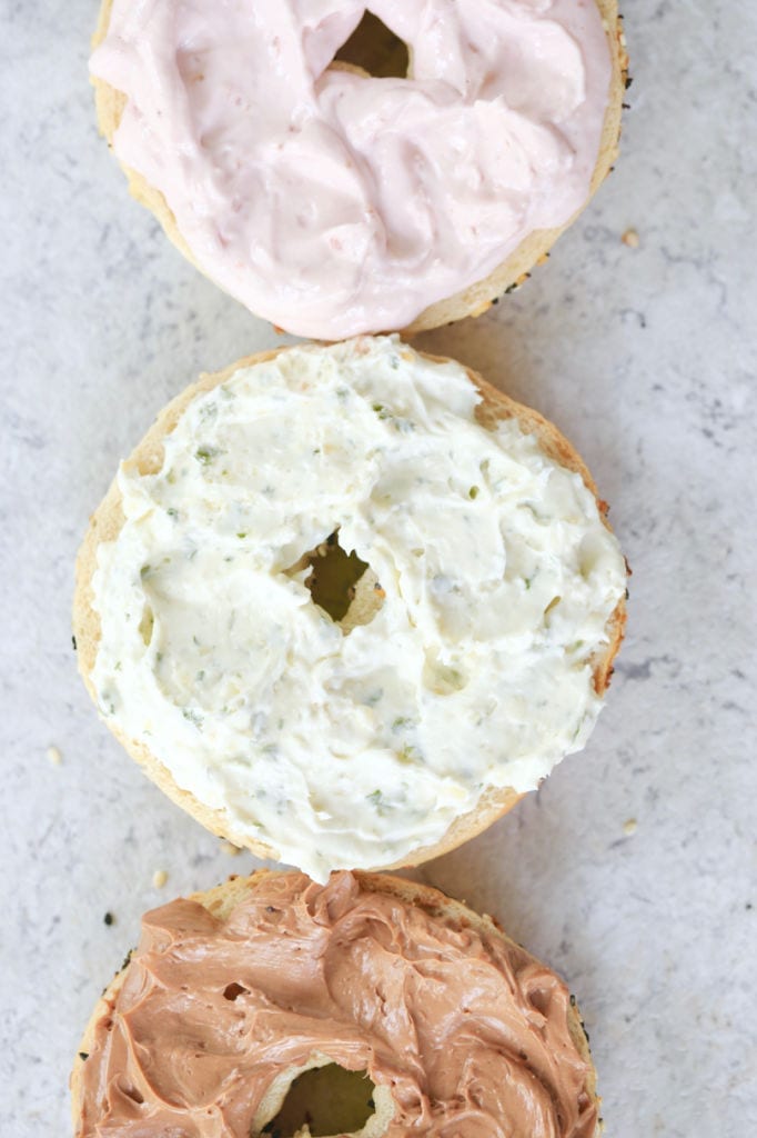 A top-down view of three bagels, each with a different homemade cream cheese flavor: strawberry, garlic & herb, and chocolate.