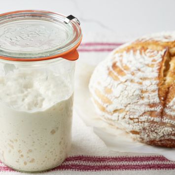 A jar of bubbly sourdough starter with a loaf of golden, crusty sourdough bread.