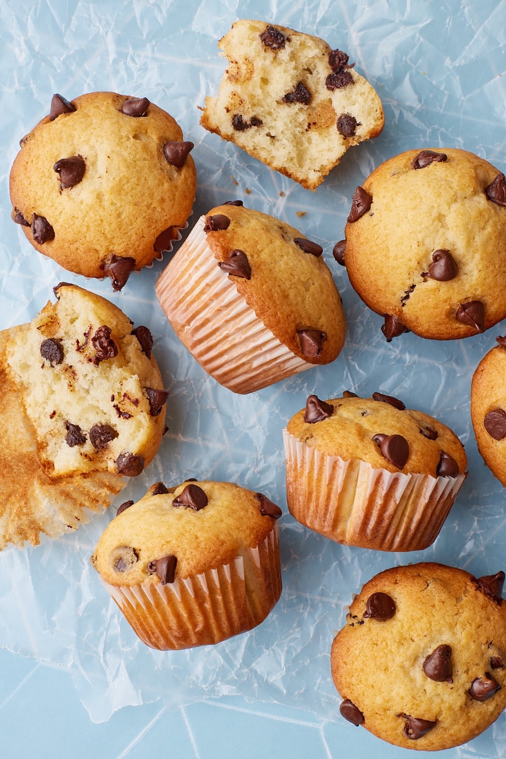Chocolate Chip Muffins7 Overhead view of homemade chocolate chip muffins with golden tops, scattered chocolate chips, and one muffin broken open to show a soft, fluffy crumb filled with chocolate chips.