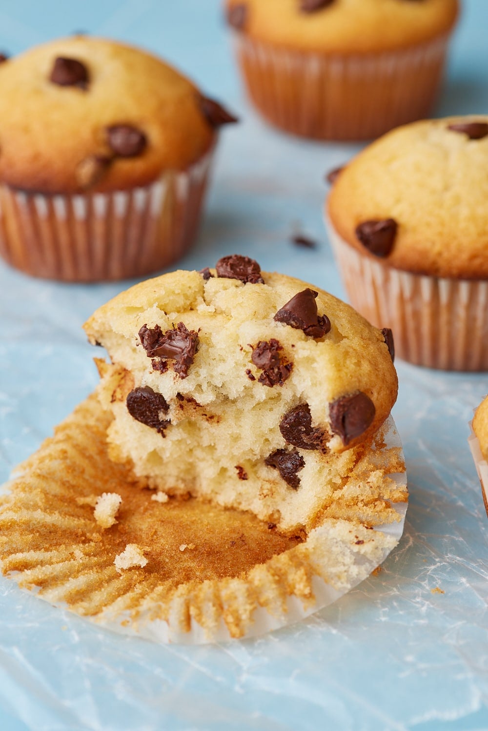 Chocolate Chip Muffins9 A close up shot at chocolate chip muffin split open, to reveal a soft, fluffy crumb packed with melted chocolate chips, with more golden muffins in the background.