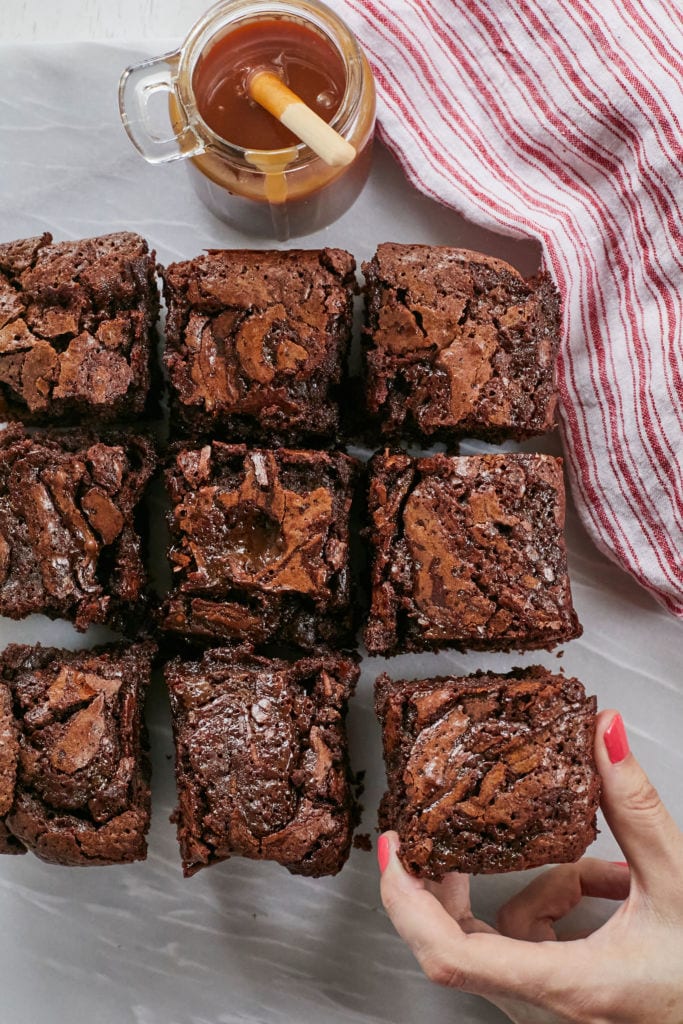 A grid of my Caramel Brownies recipe as I reach in and grab one, with a jar of caramel sauce looking on.