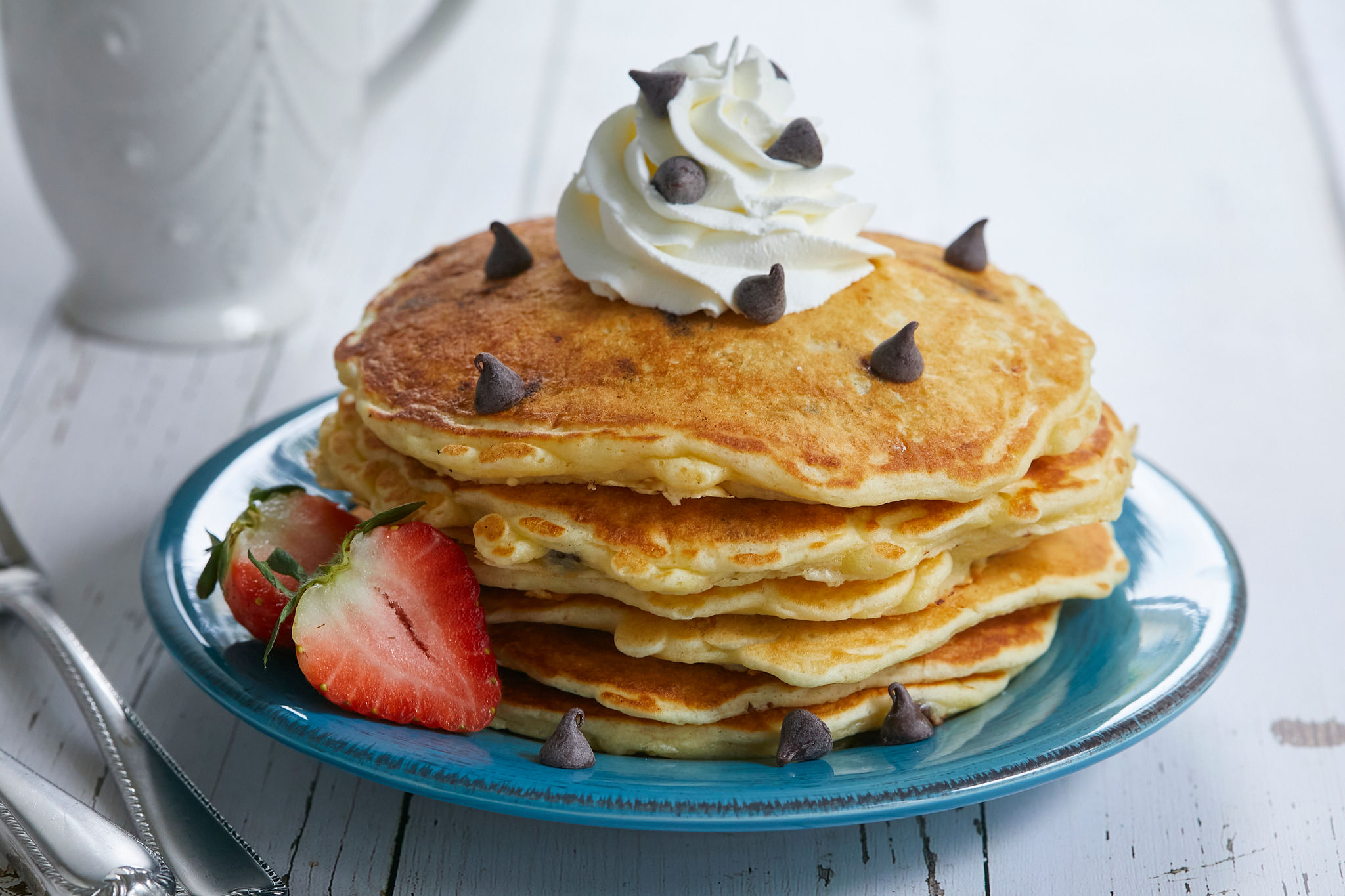 A stack of my Perfectly Sweet Chocolate Chip Pancake recipe, with chocolate chips, whipped cream, and strawberries on a blue plate.