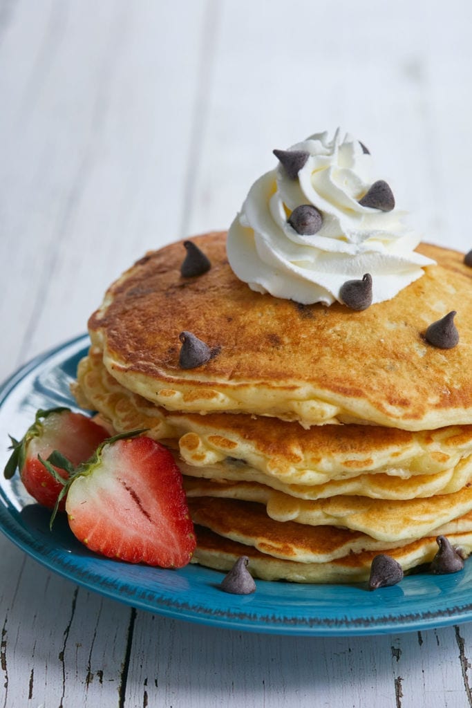 A stack of my Perfectly Sweet Chocolate Chip Pancake recipe, with chocolate chips, whipped cream, and strawberries on a blue plate on a white table.