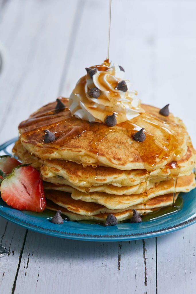 A stack of my Perfectly Sweet Chocolate Chip Pancake recipe, with chocolate chips, whipped cream, and strawberries on a blue plate being drizzled with syrup.